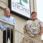 Bryn Stroyke (left) and Robb Stroyke (right) smiling on a balcony in front of their Stroyke Properties sign, introducing the article about their business.