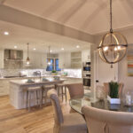 View of a modern kitchen with white shaker cabinets, light wood floors, a large island with seating, and an adjacent dining area with a glass table and stylish chandelier.