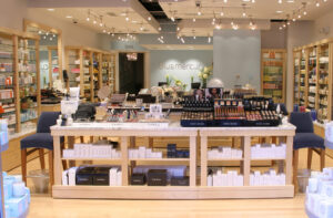 The interior of a Bluemercury store, featuring display tables laden with makeup from brands like Trish McEvoy and Bobbi Brown, surrounded by illuminated shelves stocked with various skincare and beauty products. In the background, a light blue wall shows the Bluemercury logo and a sign for the 'SPA' area, illustrating the store's dual retail and treatment offerings.