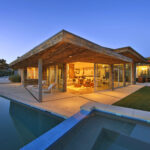 Exterior view of a modern luxury home at dusk, showcasing a large swimming pool and hot tub in the foreground, and the brightly illuminated interior living space through expansive glass doors, emphasizing a tranquil, high-end lifestyle.