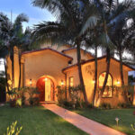 Exterior view of a Spanish Revival house with a yellow stucco facade, arched wooden front door, red tile roof, palm trees, green lawn, illuminated pathway, and a fountain, pictured at dusk.