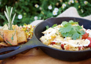 Close-up of two breakfast or brunch dishes: scrambled eggs with tomatoes and cilantro in a cast-iron skillet, and roasted potatoes with rosemary, set on a wooden table with green foliage in the background, illustrating the food offerings at Taste on Melrose.