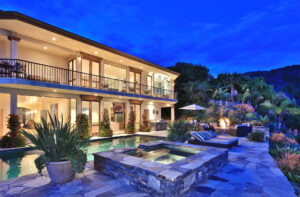 Exterior view of a luxurious multi-story home at twilight, showcasing a swimming pool, stone hot tub, fire pit lounge, and an outdoor kitchen. The property features extensive landscaping and illuminated interiors, emphasizing its grandeur and outdoor amenities.