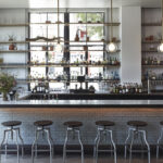 A wide shot of a contemporary bar interior by Montalba Architects, featuring a white marble counter, six metal bar stools, and illuminated shelves filled with bottles, glassware, and decorative plants.