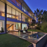 Exterior view of the Caverhill Residence by Zoltan Pali at dusk, showcasing its multi-story glass facade, illuminated interiors, and luxurious patio with a fire pit and pool.