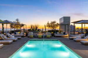 An illuminated rooftop swimming pool at dusk surrounded by modern outdoor sofas, chaise lounges, and lush desert landscaping, with the Pacific Ocean and Santa Monica cityscape visible under a colorful sunset sky, highlighting the luxury amenities of The Park Santa Monica.