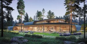 An eye-level exterior view of the Riverwave house by Kerimov Architects, showing its sweeping glass facade and organic roofline blending into a forest of tall pine trees and natural landscaping.