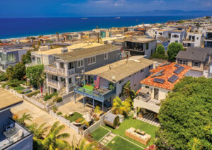 Aerial drone shot of a Manhattan Beach Walk Street neighborhood showing closely set coastal homes with the deep blue ocean in the background under a clear sky.