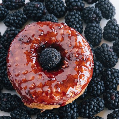 Overhead view of a glossy, berry-glazed doughnut with a single fresh blackberry in its hole, surrounded by many loose blackberries, representing Sidecar Doughnuts' handcrafted treats.