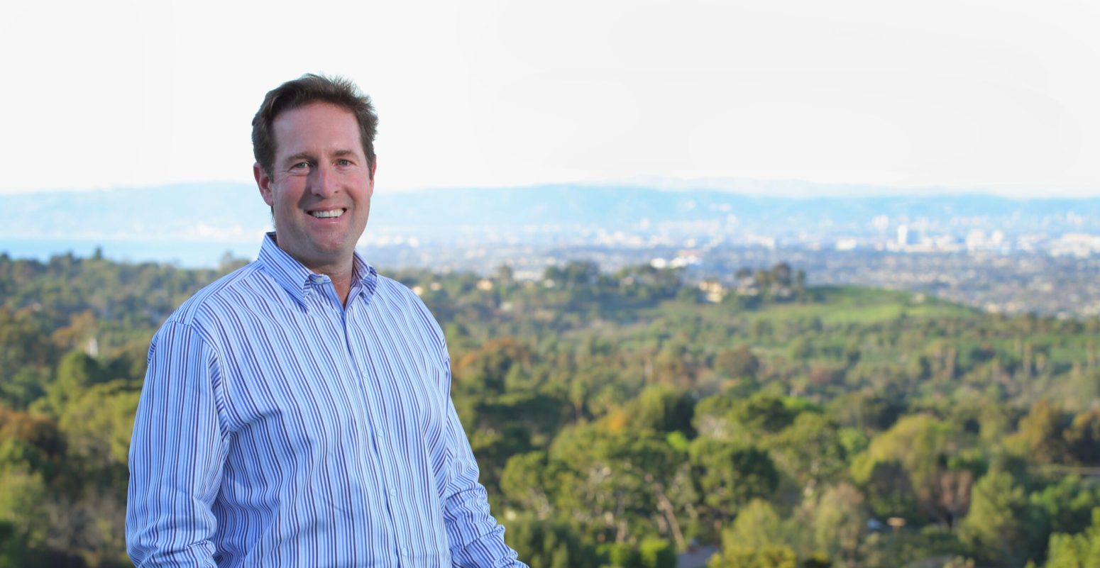 Portrait of Chris Adlam, a realtor with Vista Sotheby's International Realty, smiling with a scenic South Bay and Palos Verdes Peninsula cityscape in the background.