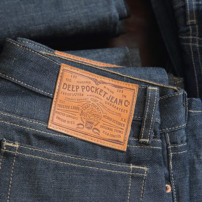 Close-up of Deep Pocket Jean Company's brown leather patch stitched onto the back of dark indigo denim jeans, part of a gallery showcasing the brand.