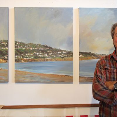 Jeff Dion, owner of Dion Gallery, stands smiling with crossed arms in front of three large, light-toned seascape oil paintings depicting a beach, ocean, and hilly coastline with houses.