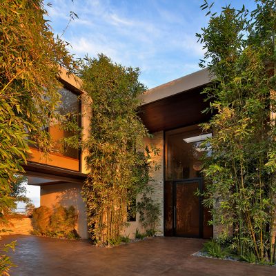 Exterior view of the Raju Chhabria-designed modern Hermosa Beach home with a grand entrance, tall glass doors, and vibrant bamboo plants under a warm sky, illustrating the article's subject.