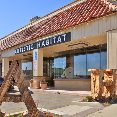 The exterior of the Artistic Habitat showroom with a terracotta roof. Large windows display interior items, while two abstract wooden sculptures resembling 'A' and 'H' stand outside, marking the entrance.