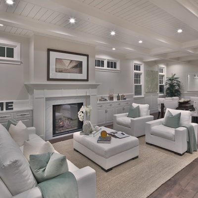 Interior of a grand living room in a Nantucket-style home, with white sofas, armchairs, and ottoman, a marble fireplace, and a dining area in the background.