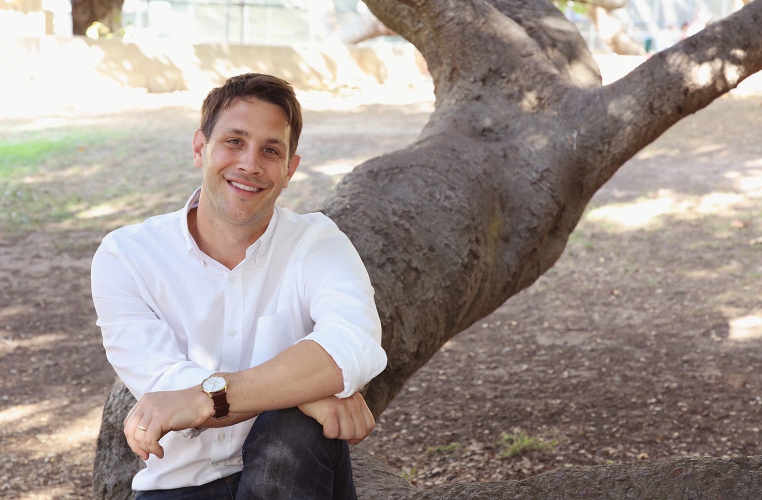 Mark Kitching, a real estate agent, sits smiling casually on a tree trunk outdoors in a white shirt and dark pants, serving as his interview portrait.