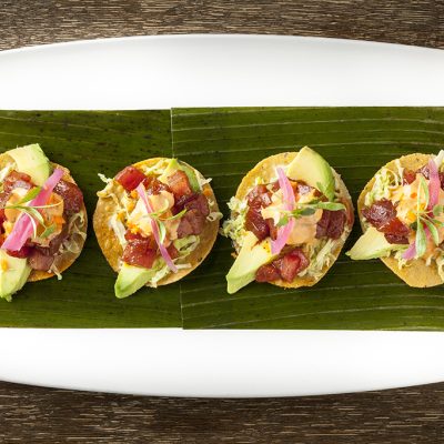 Four gourmet tuna tostadas from Rick Bayless's Red O restaurant, served on banana leaves on a white platter. The tostadas feature diced tuna, avocado, cabbage, and pickled onions.