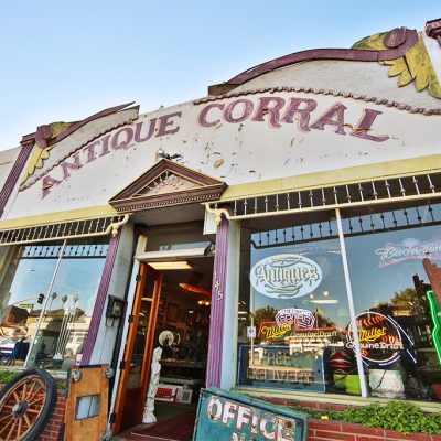 The vibrant, eclectic exterior of Antique Corral, featuring the store's name on a peeling facade, display windows filled with neon signs, and antiques like a wagon wheel visible outside.