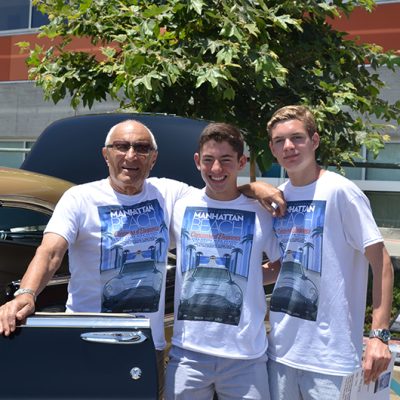 Three smiling men, including student co-founders Connor Wohl and Aidan Nesicolaci, pose with a classic gold and dark blue car at the Manhattan Beach Festival of Motoring.