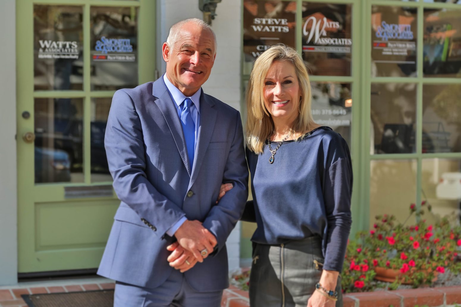 Steve and Ceci Watts, a smiling real estate agent couple, standing arm-in-arm in front of their RE/MAX Estate Properties office window, introducing the featured duo.