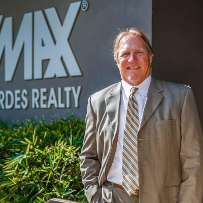 Kevin Moen, a realtor in a suit and tie, smiles in front of the "RE/MAX Palos Verdes Realty" sign, representing RE/MAX Estate Properties.