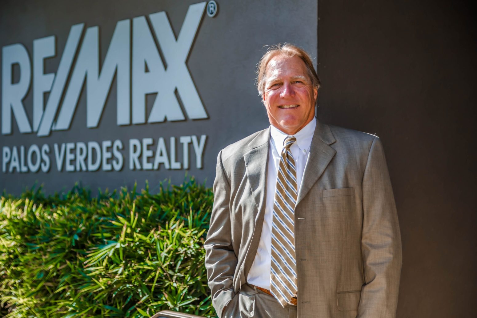 Kevin Moen, a realtor in a suit and tie, smiles in front of the "RE/MAX Palos Verdes Realty" sign, representing RE/MAX Estate Properties.