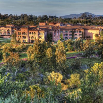 An aerial view of the L'Auberge Del Mar luxury resort, featuring a multi-story building with terracotta roofs, surrounded by vibrant green trees and manicured gardens.