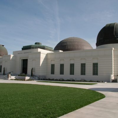 The majestic white Art Deco exterior of Griffith Observatory with its iconic domes and central entrance, seen from the lush green lawn on a clear day.