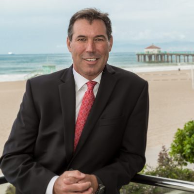Portrait of James Sanders, owner of RE/MAX Estate Properties, wearing a suit and smiling, with the Manhattan Beach Pier and ocean in the background. This image introduces the featured real estate broker.