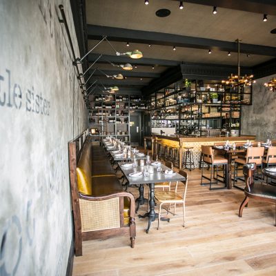 Interior of Little Sister restaurant in Manhattan Beach, showing dining tables, banquette seating, bar area, and unique decor with "little sister" stenciled on the wall.