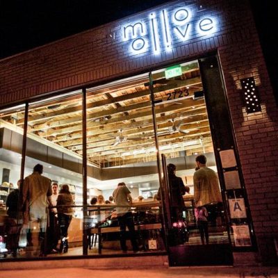 The brightly lit exterior of Milo & Olive restaurant at night, showing its neon sign, brick facade, and guests dining inside through large glass windows. This image showcases the inviting atmosphere of the featured establishment.