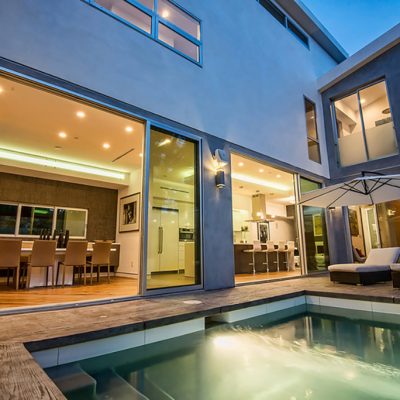 A view of a modern two-story luxury home exterior at dusk, featuring a swimming pool and patio with lounge chairs. Large glass doors reveal an illuminated dining room and kitchen, showcasing contemporary home staging.