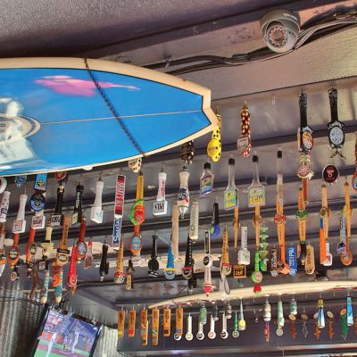 A bar ceiling densely covered with hundreds of unique, colorful beer tap handles and two blue surfboards, one clearly marked 'BUD LIGHT.'