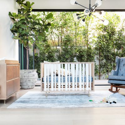 A bright, modern nursery room featuring a light wood crib with white slats, a matching changing dresser, a blue upholstered rocking chair, and a large potted plant, all illuminated by natural light from a window overlooking green foliage.