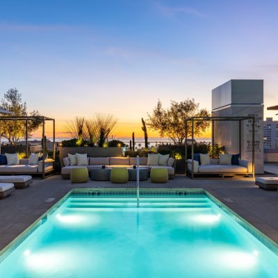 An illuminated rooftop swimming pool at dusk surrounded by modern outdoor sofas, chaise lounges, and lush desert landscaping, with the Pacific Ocean and Santa Monica cityscape visible under a colorful sunset sky, highlighting the luxury amenities of The Park Santa Monica.