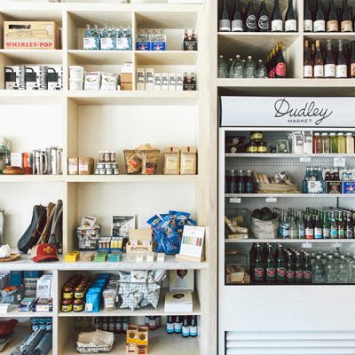 Interior view of Dudley Market's retail section, showing floor-to-ceiling shelves stocked with packaged artisanal foods, gourmet items, and kitchen goods, alongside a large white refrigerator filled with bottled beverages and fresh produce, all illustrating the market's offerings.