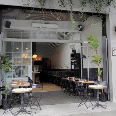 Wide view of Hinterland restaurant's open entrance on Main Street, featuring outdoor patio dining tables, potted plants, and the inviting, minimalist interior beyond.