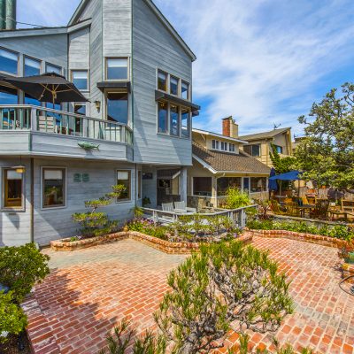 Exterior view of a gray Craftsman-inspired home with a large brick patio, lush landscaping, and an upper balcony with an umbrella, illustrating the Hermosa Beach walk street residence.