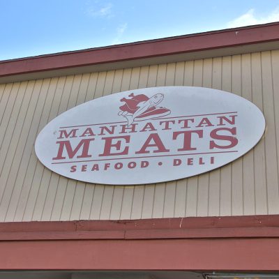 The exterior sign for "Manhattan Meats Seafood • Deli" against a beige building facade and blue sky. The white oval sign features red text, with illustrations of a chicken and a fish above the business name.