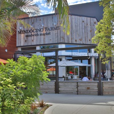 Exterior view of the new Mendocino Farms restaurant in El Segundo, California, showcasing its wooden facade, large windows, and a bustling outdoor patio area with diners.