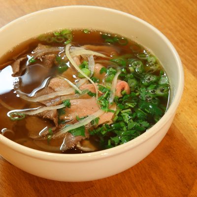 A steaming bowl of Vietnamese beef pho with thinly sliced rare beef, white onions, green onions, and fresh cilantro, featured in the article about Pho Show restaurant.