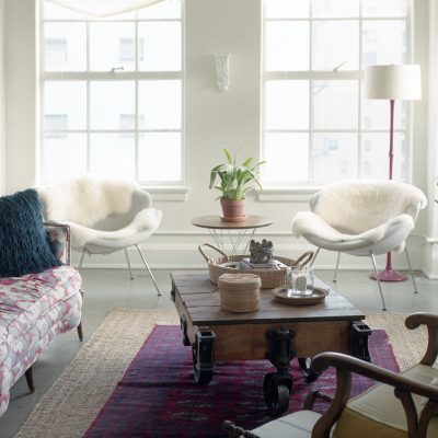 An eclectic living room designed by Ruth De Jong, with a floral sofa, two white chairs with sheepskin, a rustic wooden coffee table on wheels, potted plants, and bright windows.