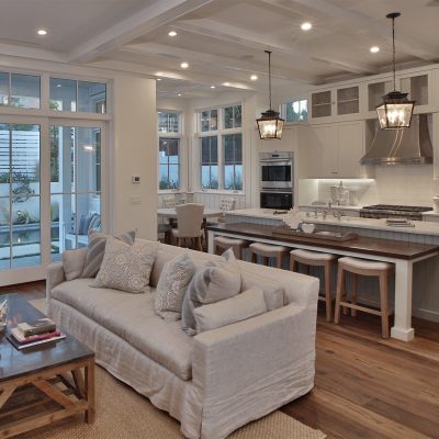 An open-plan modern farmhouse interior showing a living room with a slipcover sofa, a kitchen with a large island and bar seating, and a dining nook, all connected to an outdoor patio via glass doors.