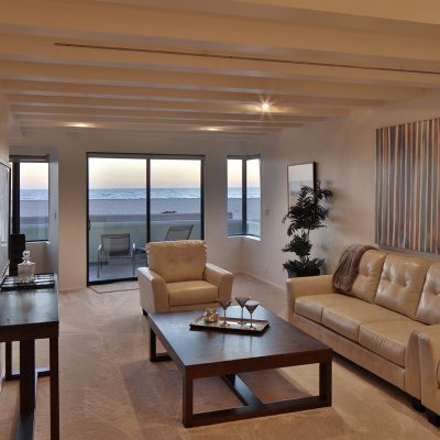 Contemporary living room with light leather sofa and armchair, dark coffee table, and large windows revealing a sandy beach and ocean view at sunset. This image showcases the beachfront home featured in the article.