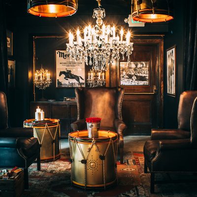 Interior of Timothy Oulton's Blue Room redesign at LAAC, showing three dark leather armchairs, two drum-shaped side tables with candles and roses, and sparkling chandeliers.