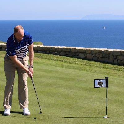 A male golfer in a striped polo shirt putts on a lush green at Trump National Golf Club, with the deep blue Pacific Ocean and a distant sailboat in the background, illustrating the course's stunning ocean views.