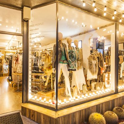 Exterior of The Urban Clothes Horse boutique, showcasing window displays with bohemian and cowboy-style outfits on mannequins, a carousel horse, and string lights. Cacti line the building facade.